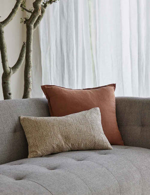 The Kisha natural-toned lumbar throw pillow sits on a gray linen sofa with a square orange throw pillow