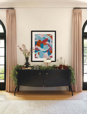 The Anabella black wood console table with silver drawer pulls sits underneath an abstract multicolored wall art with olive garlands and serveware sitting atop it.