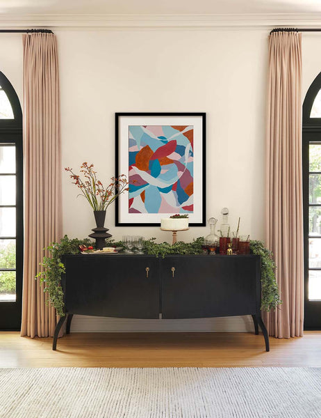 | The Anabella black wood console table with silver drawer pulls sits underneath an abstract multicolored wall art with olive garlands and serveware sitting atop it.