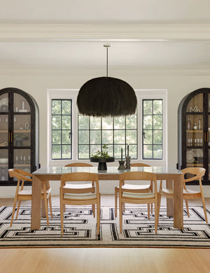 The Ida natural teak wood dining chair sits in a bright dining room surrounding a natural wood dining table atop a geometric rug underneath a fluffy chandelier with black-framed windows in the background.