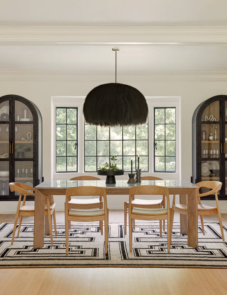 | The Paige black plush pendant light with palm tree fibers sits in a symmetrical dining room with two tall wooden sideboards, a black and beige geometric rug, and natural wooden furniture