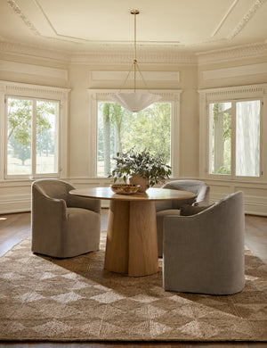 The Pau oak veneer round dining table sits atop a diamond patterned jute rug surrounded by three gray linen chairs
