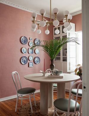 The Grasscloth rose pink tan solid wallpaper is in a breakfast nook with a circular dining table and a plate wall display