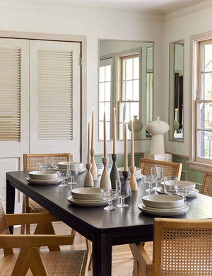 The Reese black mango wood rectangular dining table sits in a dining room surrounded by woven dining chairs with wooden candles and white dinnerware on its surface.