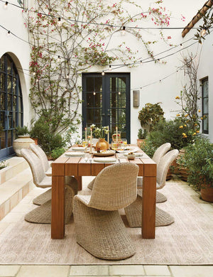 The Manila wicker weave beige indoor and outdoor dining chair sits in an outdoor dining space surrounding a fully set wooden dining table all atop a natural patterned rug
