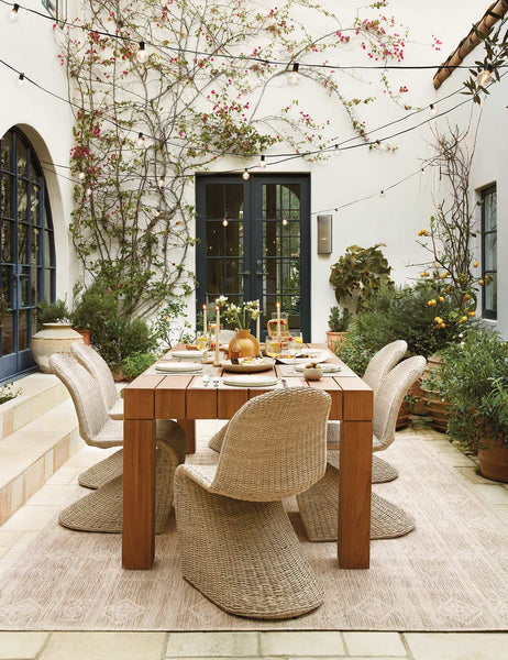 #color::natural | The Manila wicker weave beige indoor and outdoor dining chair sits in an outdoor dining space surrounding a fully set wooden dining table all atop a natural patterned rug