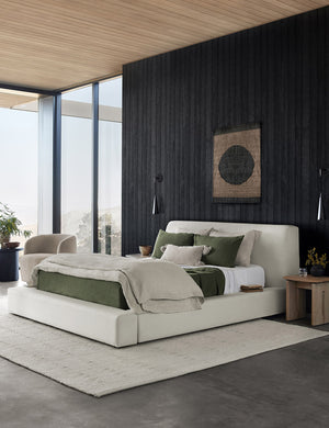 The Clayton gray upholstered platform bed sits in a bedroom with black wood paneling on the walls, olive colored bedding, and a white textured plush rug.