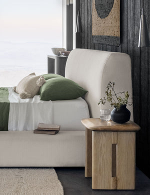 The Henrik light wood stool sits next to a natural linen framed bed with a black vase and water glass atop it