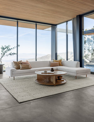 Dolan wool ivory rug lays under a circular wooden coffee table and ivory sofa in a living room with floor to ceiling windows