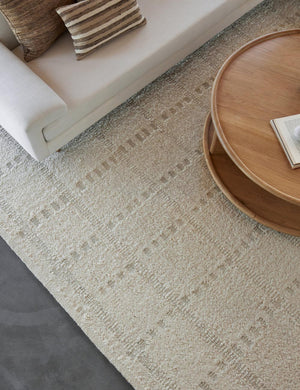 Dolan wool ivory rug with a subtle hashed motif pattern lays under a circular wooden coffee table and ivory sofa