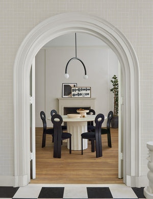 The Whit black wood sculptural dining chair by sarah sherman samuel sits in a dining room surrounding a white circular dining table with a white fireplace in the background.