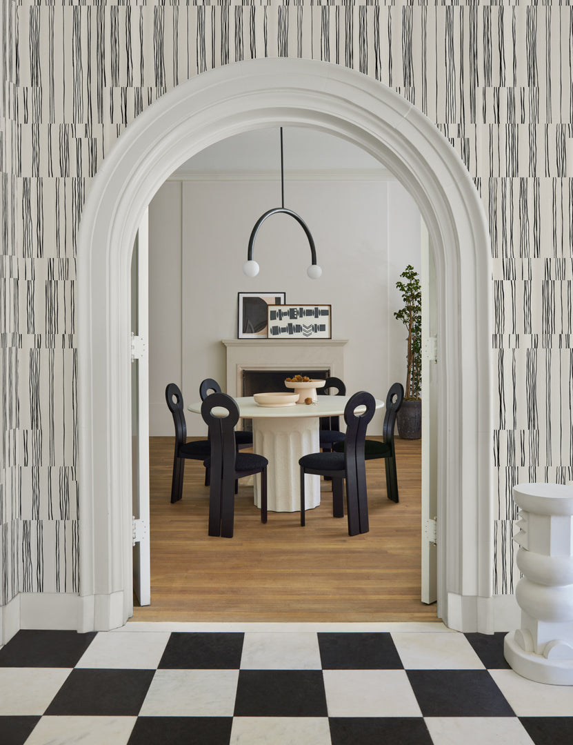 | The Broken Stripe black and white Wallpaper by Sarah Sherman Samuel featuring offset organic stripes surrounds an entryway to a dining room featuring a white round pedestal dining table and sculptural black wooden dining chairs