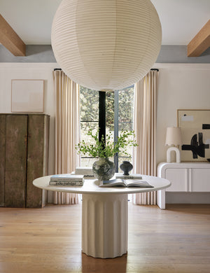 The Doric white round dining table sits underneath a large paper-lantern chandelier with a vase and stacks of books atop it