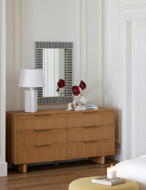 The Hillard 6-drawer dresser sits in the corner of a room with a sculptural lamp and a black and white striped mirror above it