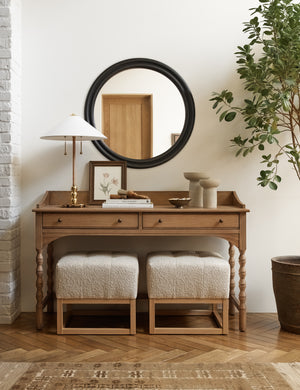 Two Taupe Boucle Grasmere Ottomans sit in a room beneath an antique desk, a table lamp, and a black framed circular mirror
