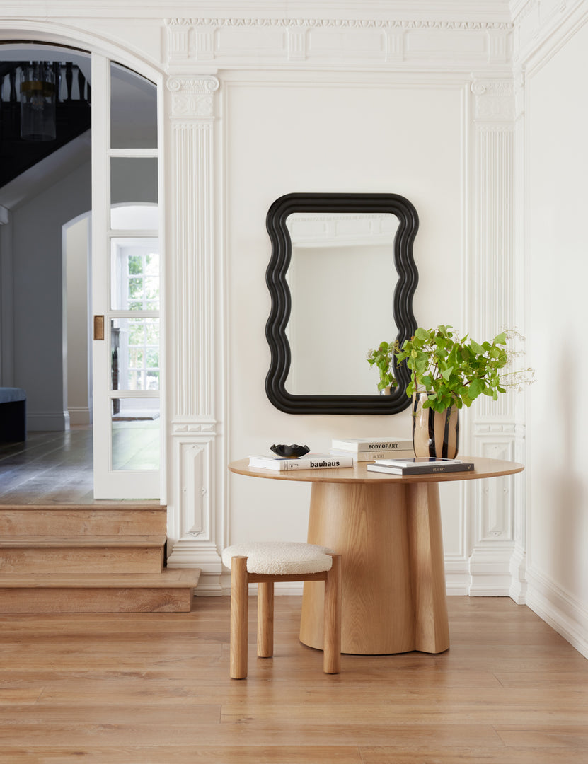 #size::large | The  | Wendolyn wavy thick-framed black wall mirror hangs on a white paneled wall over a round light wood table with books and a vase with greenery in it. | The Wendolyn large mirror hangs next to an arched entryway above a round table with a boucle stool under it