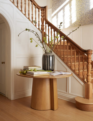 The Pau oak veneer round dining table sits next to a stair case with stacks of books and a large vase sitting atop it
