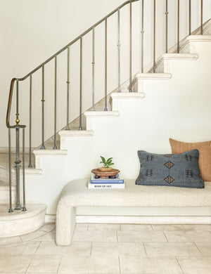 The Tate cream boucle upholstered bench sits in front of a white stone staircase with two throw pillows and a stack of books atop it.