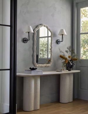 The Anastasia mirror sits atop a white sideboard with pedestal legs next to a window