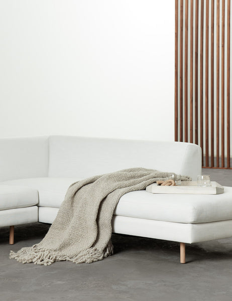 | The Goleta gray chunky wool knit throw blanket with tasseled ends lays a linen sectional sofa next to a whitewashed wooden tray