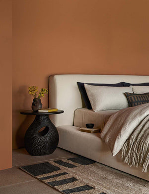 Corso black mango wood side table with a book and vase atop it sits next to a natural linen framed bed and a pattered rug
