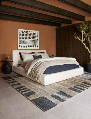 The Clayton gray upholstered platform bed sits in a bedroom with an orange and wood paneled accent wall, a polka-dotted wall art, and sculptural nightstand