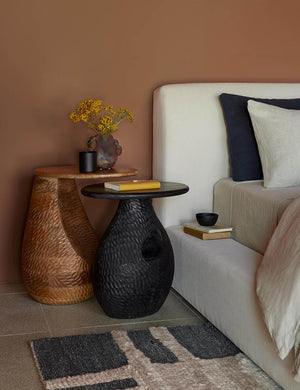The Gem mango wood side table is nested above a black side table next to a white linen bed with navy and natural linens