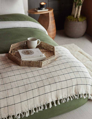 The Dani Cotton tattersall black and white Bed Cover by House No. 23 with handcrafted tasseled ends lays at the edge of a bed with olive green linens in a bedroom with a jute tray sitting atop it