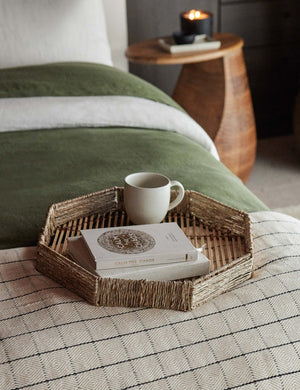 One woven geometric bamboo murai tray sits in a bedroom atop a bed with green velvet and patterned bedding with a mug and stack of books placed within it