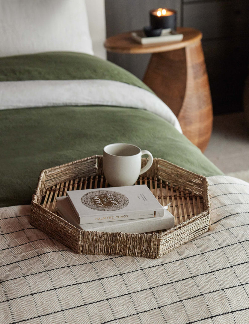| One woven geometric bamboo murai tray sits in a bedroom atop a bed with green velvet and patterned bedding with a mug and stack of books placed within it