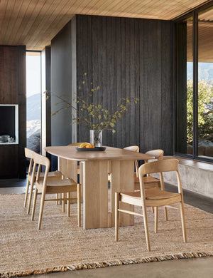 The Henrik light wood dining table sits atop a jute rug surrounded by six light wood dining chairs in front of a black wood wall