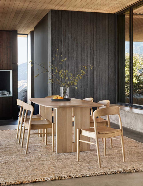 | The Henrik light wood dining table sits atop a jute rug surrounded by six light wood dining chairs in front of a black wood wall