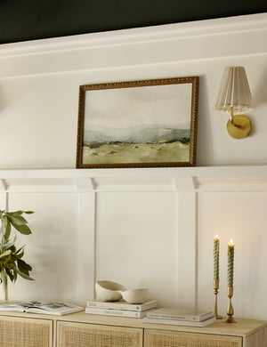 The Shepherd's Meadow Print sits on a mantel on a white accented wall next to a golden sconce and above a rattan sideboard