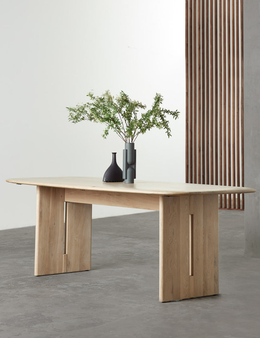 | The Henrik light wood dining table sits in a studio atop a gray floor with two sculptural black vases sitting atop it