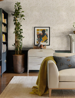 The Heritage light neutral-toned Wallpaper with a carved and etched pattern by Malene Barnett is in a living room with a white washed dresser, a colorful abstract wall art, and a natural toned sofa