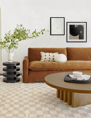 Pentwater natural wooden Round Coffee Table sits atop a white and natural checkered rug in front of a cognac velvet sofa