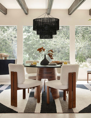 The Chavette three-tiered black jute-wrapped chandelier is hung in a dining room above a circular wooden dining table surrounded by four boucle dining chairs atop a geometric rug