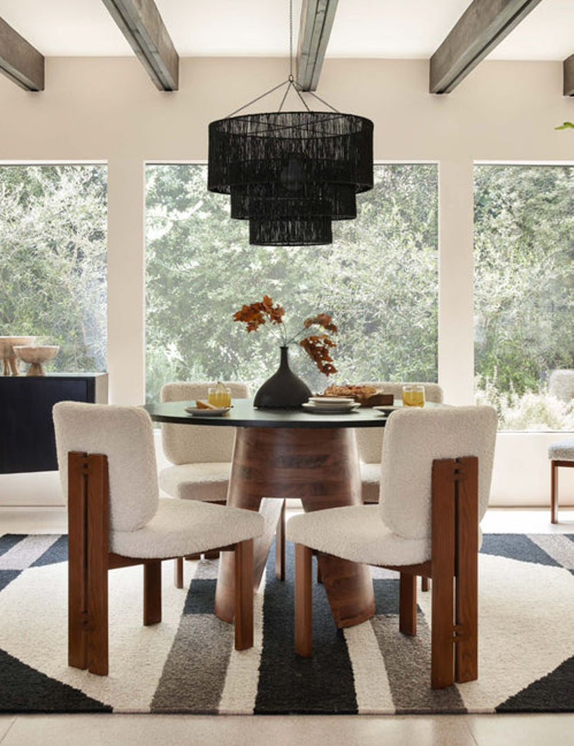 | The Chavette three-tiered black jute-wrapped chandelier is hung in a dining room above a circular wooden dining table surrounded by four boucle dining chairs atop a geometric rug