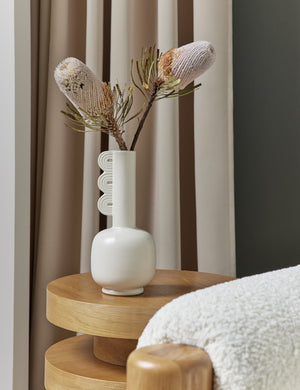 The Marguerite matte white ceramic vase with a grooved neck sits on a wooden side table with a flower inside of it
