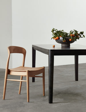 The Lakshmi terracotta bowl with antiqued black finish and woven base sits in a room on a black wooden dining table with a natural wooden ida dining chair
