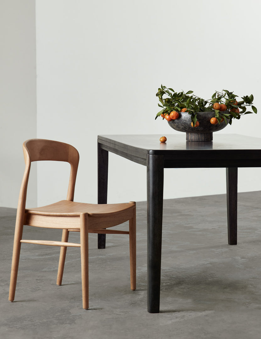 | The Lakshmi terracotta bowl with antiqued black finish and woven base sits in a room on a black wooden dining table with a natural wooden ida dining chair