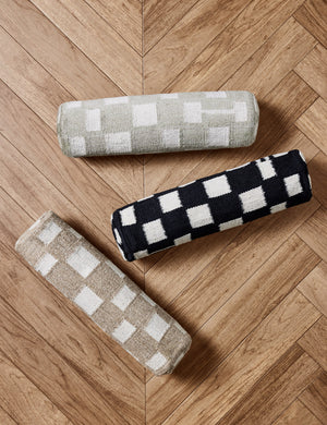 The Irregular Checkerboard Bolster Pillow by Sarah Sherman Samuel in black, taupe, and khaki green lay together on a chevron wooden floor