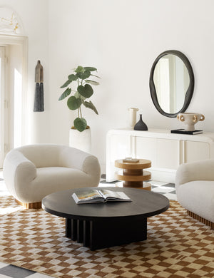 The Doreen black organic round mirror is hung in a living room with a round black dining table, two white boucle accent chairs, and an ochre and white checkered rug