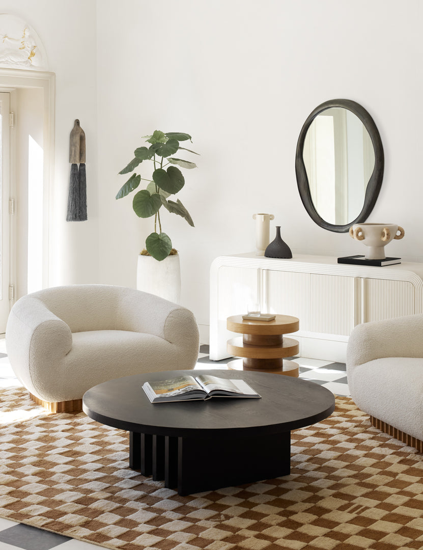 #color::black | The Doreen black organic round mirror is hung in a living room with a round black dining table, two white boucle accent chairs, and an ochre and white checkered rug