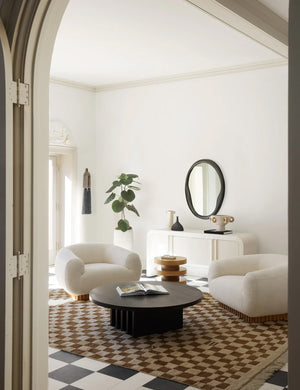 The Irregular ochre checkerboard rug by Sarah Sherman Samuel sits in a living room with a black round coffee table, white boucle rounded accent chairs, all sitting atop a checkerboard floor.