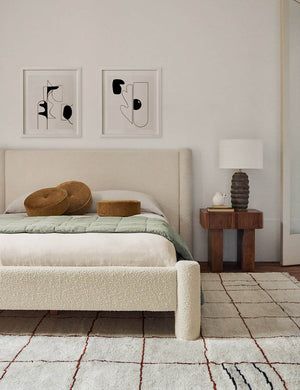 Ivory Boucle Hyvaa Bed sits in a bedroom with a grid patterned rug, two abstract walls arts hung above it, and a wooden nightstand