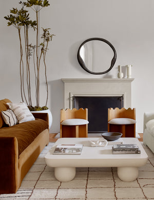 The Doreen black organic round mirror is hung in a living room with a rounded sculptural coffee table, two sculptural accent chairs, and a cognac velvet sofa