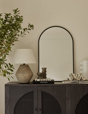 The Shashenka arched wall mirror with black frame sits top a black wooden sideboard with a sculptural lamp and decor