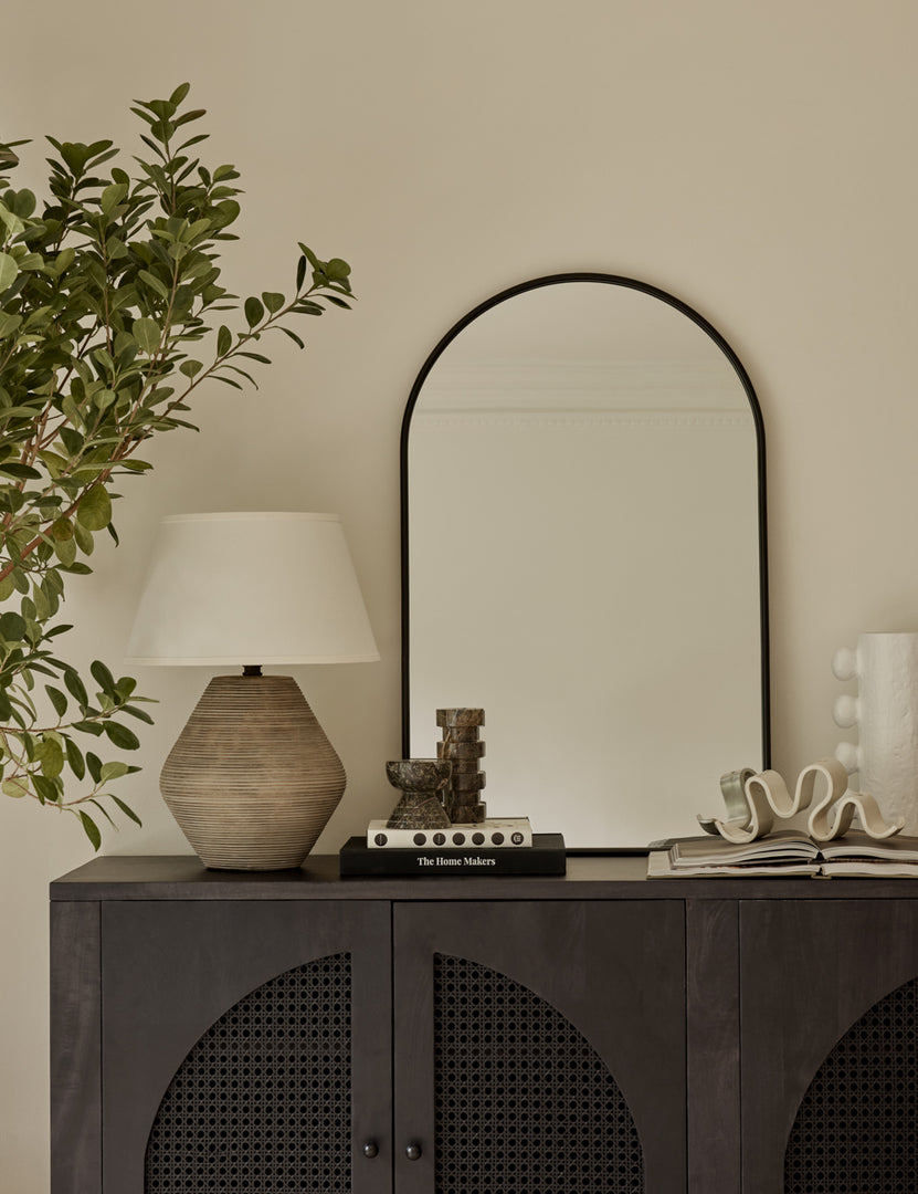 #color::black #size::38--x-24- | The Shashenka arched wall mirror with black frame sits top a black wooden sideboard with a sculptural lamp and decor