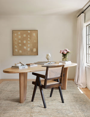 The Ismenia distressed persian rug with fringed ends lays in an office with a beige wall art, a rounded wooden desk, and a woven black framed chair